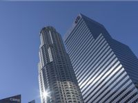 a city street in the sunlight behind two skyscrapers with cars parked along it, and a blue sky above them