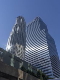 a city street in the sunlight behind two skyscrapers with cars parked along it, and a blue sky above them