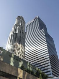 a city street in the sunlight behind two skyscrapers with cars parked along it, and a blue sky above them