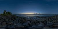 a city skyline in the distance with the moon on its horizon by water from rocks