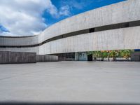 a circular concrete structure with blue sky and clouds above it in the background, there are people walking through the area