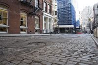 brick road surrounded by tall buildings in city area with cars parked on it's side