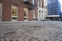 brick road surrounded by tall buildings in city area with cars parked on it's side