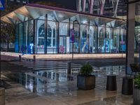 an outside area with several plants and benches next to an awning at night and light reflections on the pavement