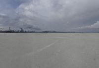 a long sandy beach has buildings in the distance and gray skies above the city and river