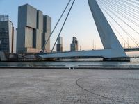 a bench sitting on the sidewalk with buildings in the background on the water side in front of a bridge