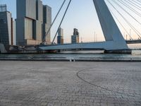 a bench sitting on the sidewalk with buildings in the background on the water side in front of a bridge