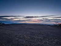 a very dark desert with some sand and bushes around it at sunset, near the horizon