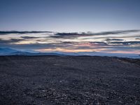 a very dark desert with some sand and bushes around it at sunset, near the horizon