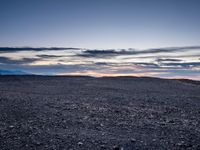 a very dark desert with some sand and bushes around it at sunset, near the horizon