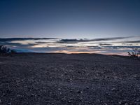 a very dark desert with some sand and bushes around it at sunset, near the horizon