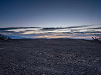 a very dark desert with some sand and bushes around it at sunset, near the horizon