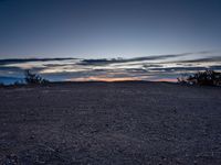 a very dark desert with some sand and bushes around it at sunset, near the horizon