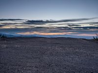 a very dark desert with some sand and bushes around it at sunset, near the horizon