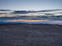 a very dark desert with some sand and bushes around it at sunset, near the horizon