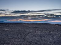 a very dark desert with some sand and bushes around it at sunset, near the horizon