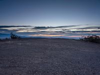 a very dark desert with some sand and bushes around it at sunset, near the horizon