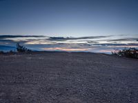 a very dark desert with some sand and bushes around it at sunset, near the horizon