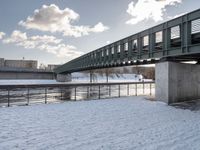 a bridge with a snow covered field and buildings in the distance on a cloudy day