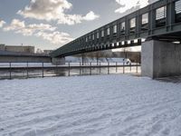 a bridge with a snow covered field and buildings in the distance on a cloudy day