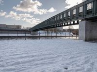 a bridge with a snow covered field and buildings in the distance on a cloudy day