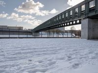 a bridge with a snow covered field and buildings in the distance on a cloudy day