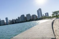 two people walk along a walkway by the water in front of city buildings and a body of