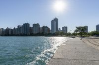 two people walk along a walkway by the water in front of city buildings and a body of