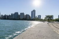 two people walk along a walkway by the water in front of city buildings and a body of