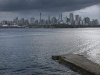 an empty bench is overlooking a large city on the water and on a cloudy day