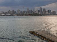 an empty bench is overlooking a large city on the water and on a cloudy day