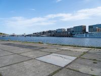 a concrete sidewalk near the water with many buildings in the back ground and a bridge over it