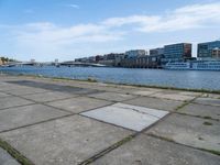 a concrete sidewalk near the water with many buildings in the back ground and a bridge over it