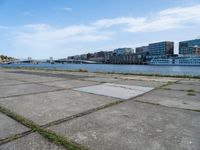 a concrete sidewalk near the water with many buildings in the back ground and a bridge over it