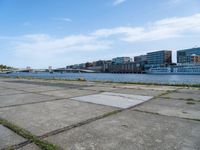 a concrete sidewalk near the water with many buildings in the back ground and a bridge over it