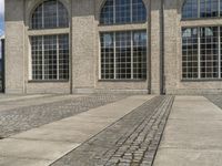 a large brick building that has some windows near by it on the street and next to a brick walkway