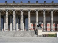 a group of pillars are in front of a building with large columns, and a sculpture is on the top of the steps