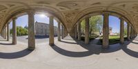 a 360 camera lens view shows pillars and arches, in a garden in front of a stone building