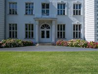 the front of a large, white building with several windows and many flower beds in the lawn