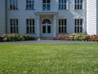 the front of a large, white building with several windows and many flower beds in the lawn