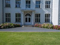 the front of a large, white building with several windows and many flower beds in the lawn