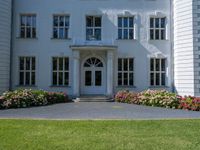 the front of a large, white building with several windows and many flower beds in the lawn