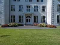 the front of a large, white building with several windows and many flower beds in the lawn