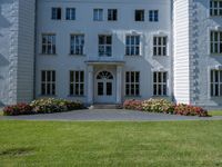 the front of a large, white building with several windows and many flower beds in the lawn