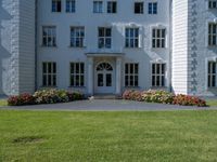 the front of a large, white building with several windows and many flower beds in the lawn