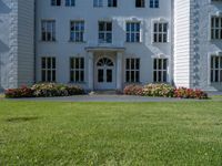 the front of a large, white building with several windows and many flower beds in the lawn