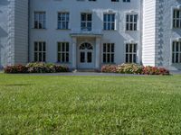 the front of a large, white building with several windows and many flower beds in the lawn