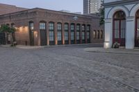 brick paved square outside a large building with double doors in the center and plants on either side of the door