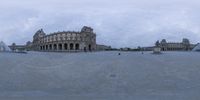 a large building sits in a square with a lot of people sitting on the grass