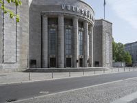 a city street next to a big building in the background, with columns and pillars on both sides of the building